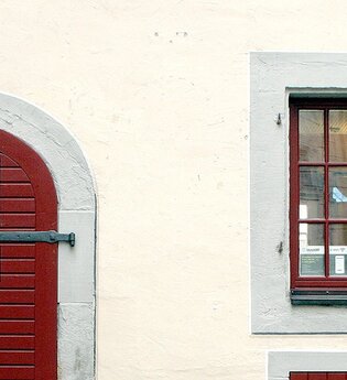 Eingangstüre zur Touristinformation aus dunkelrot gestrichenem Holz, rechts daneben zwei, dunkelrot gestrichene Sprossenfenstertüre,