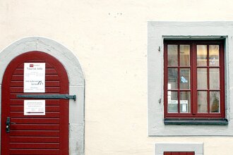Eingangstüre zur Touristinformation aus dunkelrot gestrichenem Holz, rechts daneben zwei, dunkelrot gestrichene Sprossenfenstertüre,