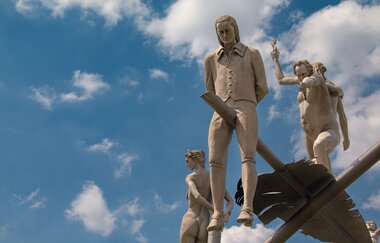 Am Ortseingang von Lauffen am Neckar erhebt sich das eindrucksvolle Kunstwerk „Hölderlin im Kreisverkehr“ des Bildhauers Peter Lenk. Auf einem geschwungenen „H“ balanciert eine überdimensionale Schreibfeder, auf der Friedrich Hölderlin als Kind und als ju
