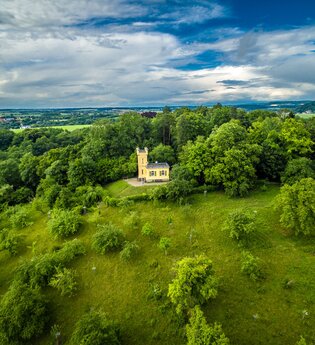 Eine kleine Villa mit Turm steht auf einem grünen Hügel, umgeben von grünen Bäumen.