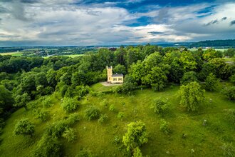 Eine kleine Villa mit Turm steht auf einem grünen Hügel, umgeben von grünen Bäumen.