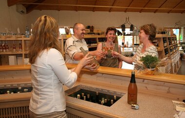 Vier Personen stehen an der Theke und halten Gläser mit Wein und Sekt. Eine Person schenkt mit einer Flasche ein.