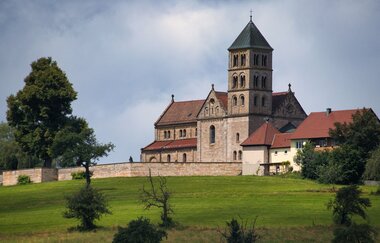 Romanische Kirche auf Hügel umgeben von Mauer. Grüne Wiesen und Bäume.